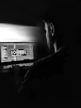 A person is focused intently on a laptop in a dark setting. The screen displays complex software with white text and graphics, suggesting a technical or creative task. The lighting is dim, with the glow from the laptop being the primary light source, casting shadows.