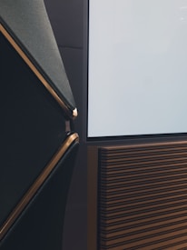 A modern audio setup featuring a stylish speaker with dark gray and gold accents. The speaker has a textured surface with horizontal lines and sits beside a flat screen or panel with a reflective surface.