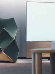 A modern interior space with a large, abstract geometric structure featuring dark green panels and gold outlines. Adjacent to it is a minimalist wooden table with rounded legs. In the background, a large white panel with a light wooden base appears to be part of the wall, suggesting a sleek, contemporary design theme.
