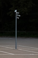 A tall metal pole stands in an empty parking lot, equipped with multiple security cameras pointed in different directions, surrounded by a background of dense, dark greenery.
