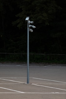 A tall metal pole stands in an empty parking lot, equipped with multiple security cameras pointed in different directions, surrounded by a background of dense, dark greenery.