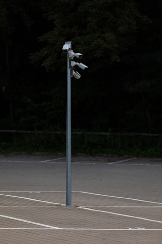 A tall metal pole stands in an empty parking lot, equipped with multiple security cameras pointed in different directions, surrounded by a background of dense, dark greenery.