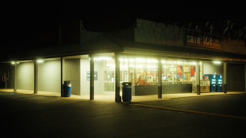 A dimly lit convenience store at night, featuring a glowing interior with fluorescent lights. The storefront has bright windows, a visible counter with products, and vending machines to the right. The atmosphere is quiet and slightly eerie due to the nighttime setting.