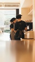 Two baristas are working behind a counter in a coffee shop, wearing black uniforms and aprons. They are focused on preparing drinks, with various coffee cups lined up on shelves beside them. The light from outside the shop casts a warm glow across the interior.