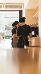 Two baristas are working behind a counter in a coffee shop, wearing black uniforms and aprons. They are focused on preparing drinks, with various coffee cups lined up on shelves beside them. The light from outside the shop casts a warm glow across the interior.