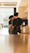 Two baristas are working behind a counter in a coffee shop, wearing black uniforms and aprons. They are focused on preparing drinks, with various coffee cups lined up on shelves beside them. The light from outside the shop casts a warm glow across the interior.