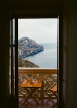 A picturesque view of a mountain and ocean landscape framed by an open balcony door. The balcony features a small wooden table and chair, and the scenery is composed of rugged cliffs extending into calm blue waters under a slightly overcast sky.