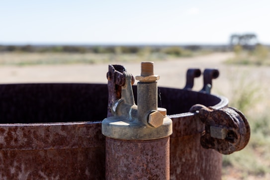 A close-up of a rusted metal container with a valve, placed outdoors. The metal appears weathered, suggesting exposure to the elements over time. In the background, there is a blurred view of a natural landscape, possibly a desert or arid region.