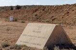 A triangular concrete sign with warning text about buried radioactive materials is situated in a barren, rocky desert landscape. The terrain is dry and rugged with sparse vegetation, and a barrel labeled 'Taranaki' is visible in the background.