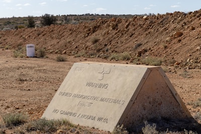 A triangular concrete sign with warning text about buried radioactive materials is situated in a barren, rocky desert landscape. The terrain is dry and rugged with sparse vegetation, and a barrel labeled 'Taranaki' is visible in the background.