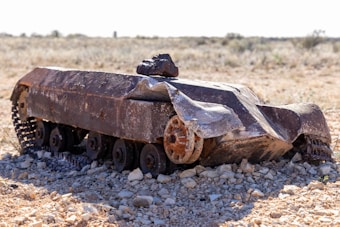 A rusted, damaged vehicle resembling a tank sits abandoned on a rocky terrain under an open, clear sky in what appears to be a desolate and arid environment.