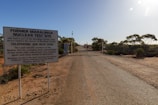 A dry and desolate landscape stretches out with a rugged road leading to a gate. Large warning signs indicate the area as the former Maralinga nuclear test site. The surrounding area is barren with sparse vegetation and eucalyptus trees. The sky is clear and bright, contributing to a stark and isolated atmosphere.