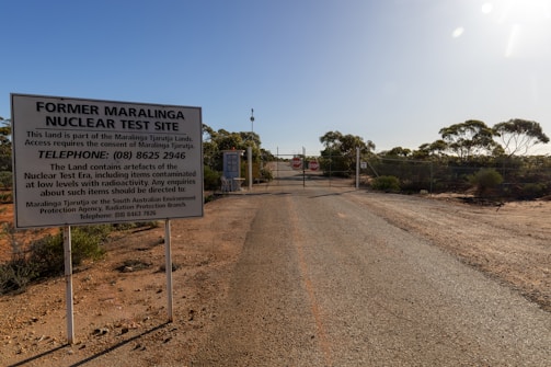 A dry and desolate landscape stretches out with a rugged road leading to a gate. Large warning signs indicate the area as the former Maralinga nuclear test site. The surrounding area is barren with sparse vegetation and eucalyptus trees. The sky is clear and bright, contributing to a stark and isolated atmosphere.