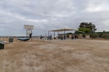 A remote fuel station with a large fuel sign and a sculpture of a whale tail. There are several fuel pumps underneath a canopy structure. The surrounding area is flat and barren, with sparse vegetation and a few trees. The sky is overcast, giving a dull appearance to the scene. A small building is adjacent to the fuel pumps, possibly a store or a rest stop.