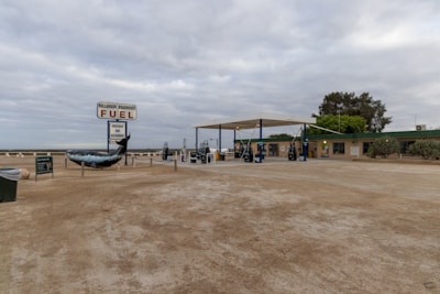 A remote fuel station with a large fuel sign and a sculpture of a whale tail. There are several fuel pumps underneath a canopy structure. The surrounding area is flat and barren, with sparse vegetation and a few trees. The sky is overcast, giving a dull appearance to the scene. A small building is adjacent to the fuel pumps, possibly a store or a rest stop.