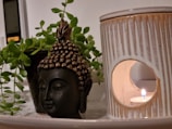 A serene setup featuring a small Buddha head sculpture placed beside a ceramic candle holder, with a lit candle casting a warm glow. Lush green leaves from a potted plant partially encircle the Buddha sculpture, creating a tranquil and harmonious arrangement.