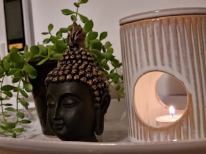 A serene setup featuring a small Buddha head sculpture placed beside a ceramic candle holder, with a lit candle casting a warm glow. Lush green leaves from a potted plant partially encircle the Buddha sculpture, creating a tranquil and harmonious arrangement.