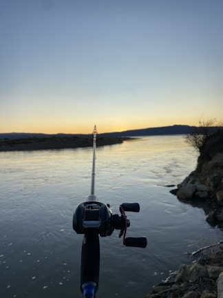 A sturdy fishing rod leaning against a calm riverbank at sunrise, with mist rising off the water.