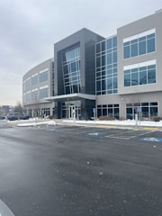 A modern office building with a glass facade and several stories. The structure features large windows and a sleek design. The surrounding area is a parking lot with a few visible cars, and patches of snow are present on the ground, indicating a cold season.