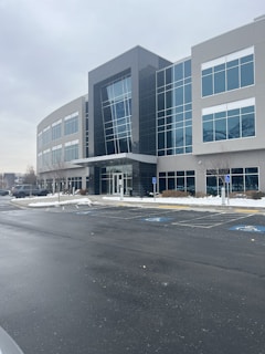 A modern office building with a glass facade and several stories. The structure features large windows and a sleek design. The surrounding area is a parking lot with a few visible cars, and patches of snow are present on the ground, indicating a cold season.