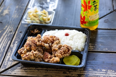 A bento box filled with pieces of crispy fried chicken, a serving of white rice topped with a small red pickled plum, and slices of yellow pickles. In the background, there is a side dish of pickled vegetables in a clear plastic tray. A bottle with green and yellow packaging featuring a maple leaf design is placed on the wooden surface where the meal is set.