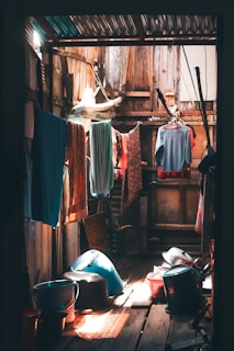A rustic wooden interior bathed in soft, warm light. Various clothes are hung on lines for drying, with colorful garments and textiles adding vividness to the space. In the foreground, there are several plastic containers and buckets, suggesting a utilitarian use of the area for laundry or storage. The atmosphere is cozy but cluttered, with light streaming through gaps in the wooden ceiling and walls, creating a play of shadows.