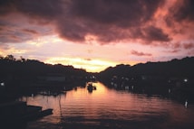 A serene waterfront scene captured at sunset, with a boat gently gliding along the reflective water surface. The sky is painted with hues of orange, pink, and purple, creating a dramatic contrast with the dark silhouette of the surrounding trees and buildings.