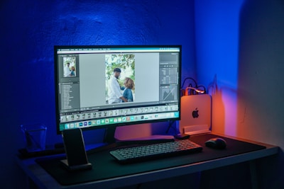 A computer setup is illuminated with blue and orange lighting. The monitor displays a photo editing software with an image of two people, possibly a couple, standing together outdoors. The desk features a keyboard, mouse, smartphone stand with a phone, and a small desktop computer with a visible Apple logo. The overall setting suggests a workspace focused on digital editing or photography.