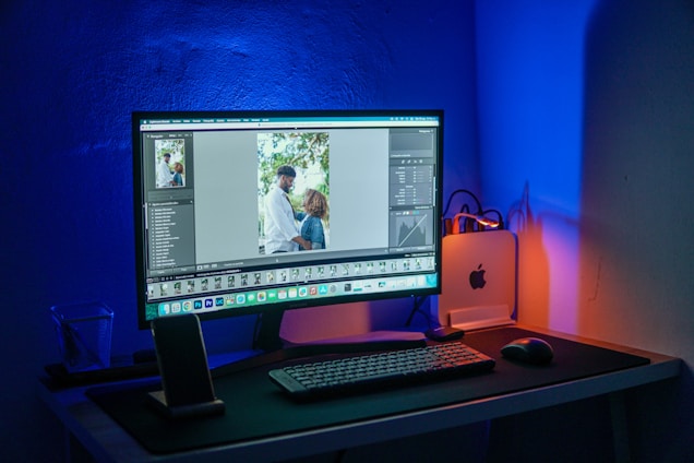 A computer setup is illuminated with blue and orange lighting. The monitor displays a photo editing software with an image of two people, possibly a couple, standing together outdoors. The desk features a keyboard, mouse, smartphone stand with a phone, and a small desktop computer with a visible Apple logo. The overall setting suggests a workspace focused on digital editing or photography.