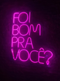 Bright neon pink text is arranged against a dark background, spelling out the phrase 'Foi bom pra voc&ecirc;?' in a vertical stack. The vivid color contrasts sharply with the dark surroundings, creating a striking and modern visual.