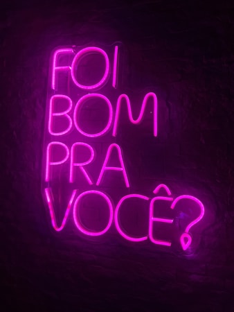 Bright neon pink text is arranged against a dark background, spelling out the phrase 'Foi bom pra voc&ecirc;?' in a vertical stack. The vivid color contrasts sharply with the dark surroundings, creating a striking and modern visual.