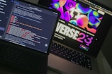 Two open laptops are placed side by side on a desk. One laptop displays a coding interface with lines of code in various colors, while the other shows a vibrant, abstract website design with bold text and colorful shapes.