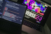 Two open laptops are placed side by side on a desk. One laptop displays a coding interface with lines of code in various colors, while the other shows a vibrant, abstract website design with bold text and colorful shapes.