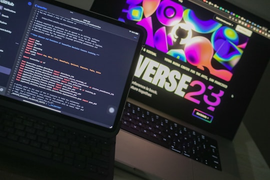 Two open laptops are placed side by side on a desk. One laptop displays a coding interface with lines of code in various colors, while the other shows a vibrant, abstract website design with bold text and colorful shapes.