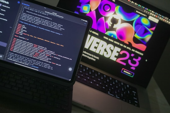Two open laptops are placed side by side on a desk. One laptop displays a coding interface with lines of code in various colors, while the other shows a vibrant, abstract website design with bold text and colorful shapes.