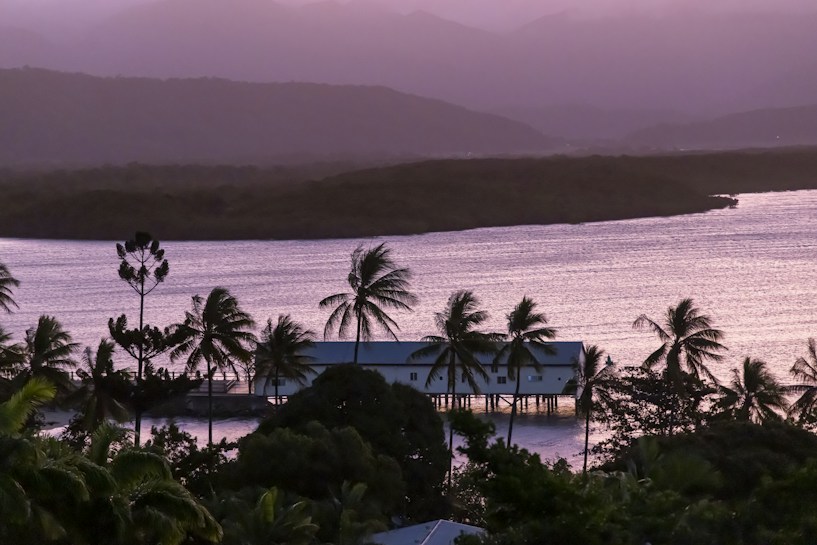 A tropical landscape showcasing a serene body of water with gentle waves glistening under soft lighting. Coconut palm trees sway along the coastline, with a stilt house sitting above the water. The background reveals misty hills under a pastel-colored sky, creating a peaceful and idyllic scenery.