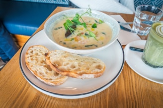 A dining table setup with a bowl of creamy soup garnished with fresh herbs, accompanied by two slices of rustic bread on a white plate. There's a glass of water, a napkin, and a knife placed on the table, along with a matcha latte in a glass on a small plate.