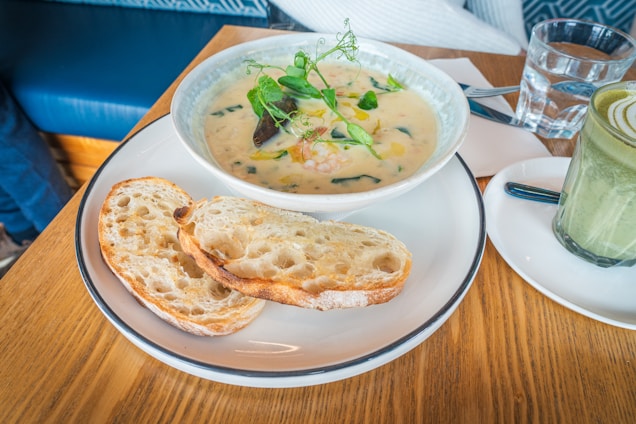 A dining table setup with a bowl of creamy soup garnished with fresh herbs, accompanied by two slices of rustic bread on a white plate. There's a glass of water, a napkin, and a knife placed on the table, along with a matcha latte in a glass on a small plate.
