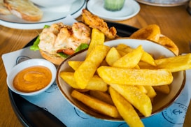 A plate of golden, crispy fries is served alongside a dipping sauce in a small bowl. Next to it, there is a burger topped with shrimp in a creamy dressing, lettuce, and tomato. Some onion rings are also visible, adding to the assortment of fried dishes. The table is set with a modern presentation, and there is a piece of toasted bread on a separate plate in the background.