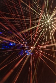 Bright, colorful laser beams radiate in various directions against a dark background, creating a dynamic and energetic composition. The beams converge towards the center and display a spectrum of colors including red, yellow, and hints of blue. The vibrant lights create a sense of movement and depth.