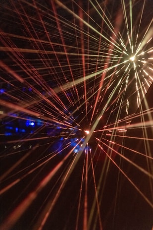 Bright, colorful laser beams radiate in various directions against a dark background, creating a dynamic and energetic composition. The beams converge towards the center and display a spectrum of colors including red, yellow, and hints of blue. The vibrant lights create a sense of movement and depth.