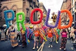 A group of people parades through a street holding large, colorful balloon letters spelling 'PROUD'. They are accompanied by vibrant costumes and balloon decorations, with large inflatable bees visible in the background. The street is lined with historical buildings with ornate facades.