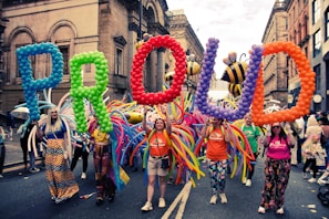 A group of people parades through a street holding large, colorful balloon letters spelling 'PROUD'. They are accompanied by vibrant costumes and balloon decorations, with large inflatable bees visible in the background. The street is lined with historical buildings with ornate facades.