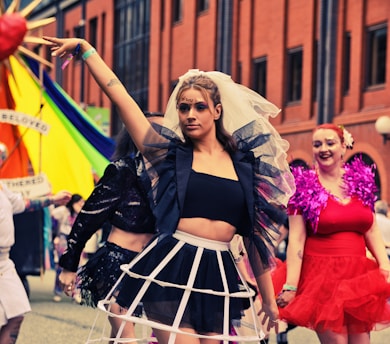 A vibrant scene with people dressed in colorful, eye-catching costumes. One person is centrally positioned wearing a distinctive black top with a white cage-style skirt and tulle on the arms, while others in the background are similarly dressed festively. The event appears to be taking place outdoors in an urban setting, with brick buildings and a rainbow-colored flag visible.