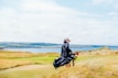 A person carrying a golf bag across a scenic golf course with a view of the ocean and distant hills under a partly cloudy sky. The landscape is composed of vibrant green grass and scattered tall grasses with a picturesque water body visible in the background.