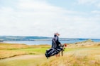 A person carrying a golf bag across a scenic golf course with a view of the ocean and distant hills under a partly cloudy sky. The landscape is composed of vibrant green grass and scattered tall grasses with a picturesque water body visible in the background.