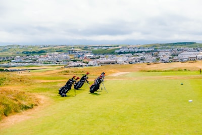 A scenic golf course stretches out under a cloudy sky with several golf bags standing on a grassy area. Behind the course, a distant town is visible, featuring numerous houses scattered over rolling green hills.
