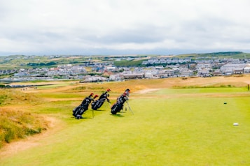 A scenic golf course stretches out under a cloudy sky with several golf bags standing on a grassy area. Behind the course, a distant town is visible, featuring numerous houses scattered over rolling green hills.