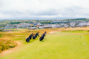 A scenic golf course stretches out under a cloudy sky with several golf bags standing on a grassy area. Behind the course, a distant town is visible, featuring numerous houses scattered over rolling green hills.