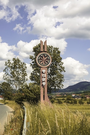 A tall signpost with an unusual design, featuring geometric symbols and text, stands by a roadside amidst tall grass and wildflowers. Two trees slightly obscure the sign, and in the distance, hills and a village are visible under a sky filled with scattered clouds.
